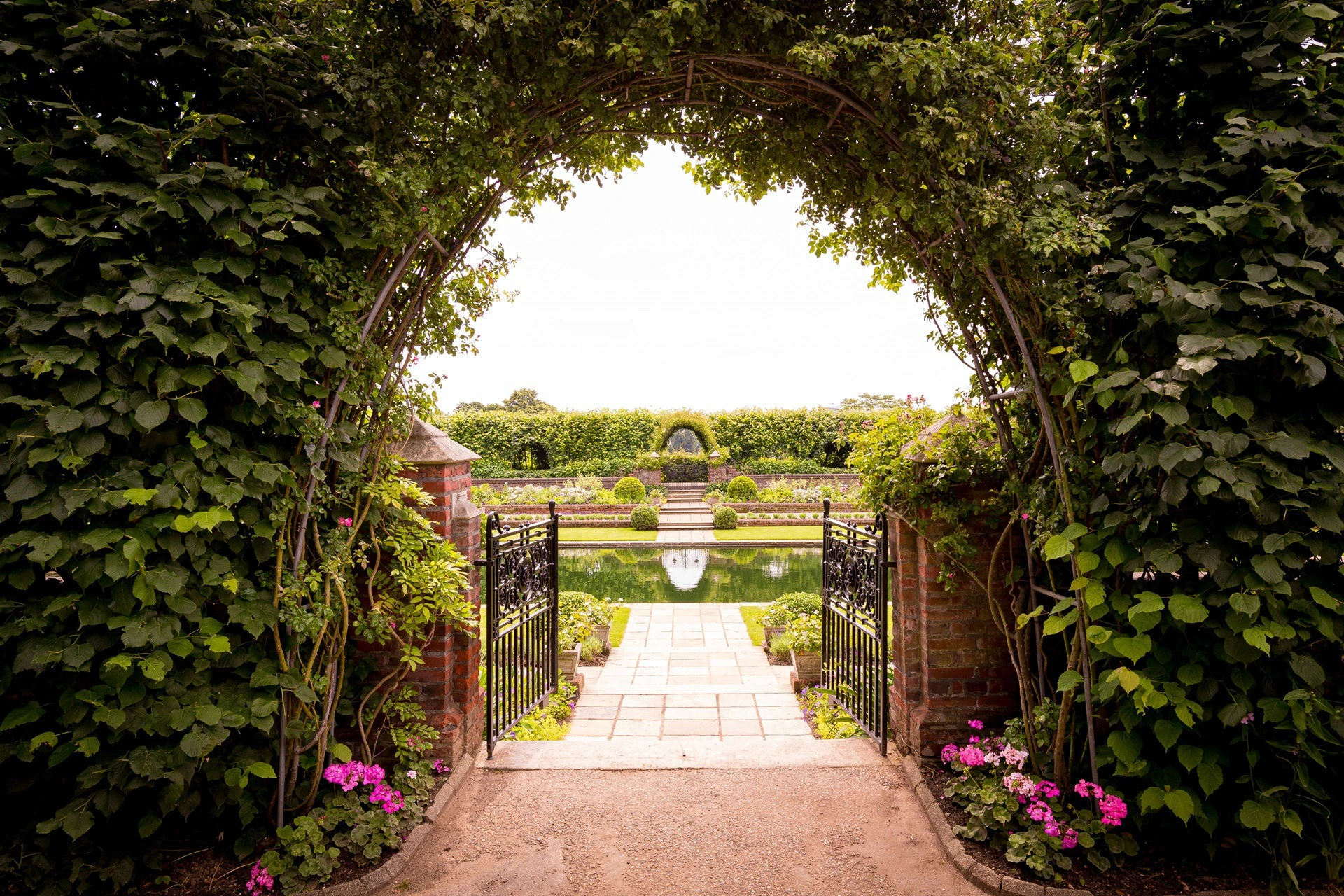 Wrought iron gate opens to a brick path leading to a reflective pond, with manicured gardens, flowers, and an arched trellis.