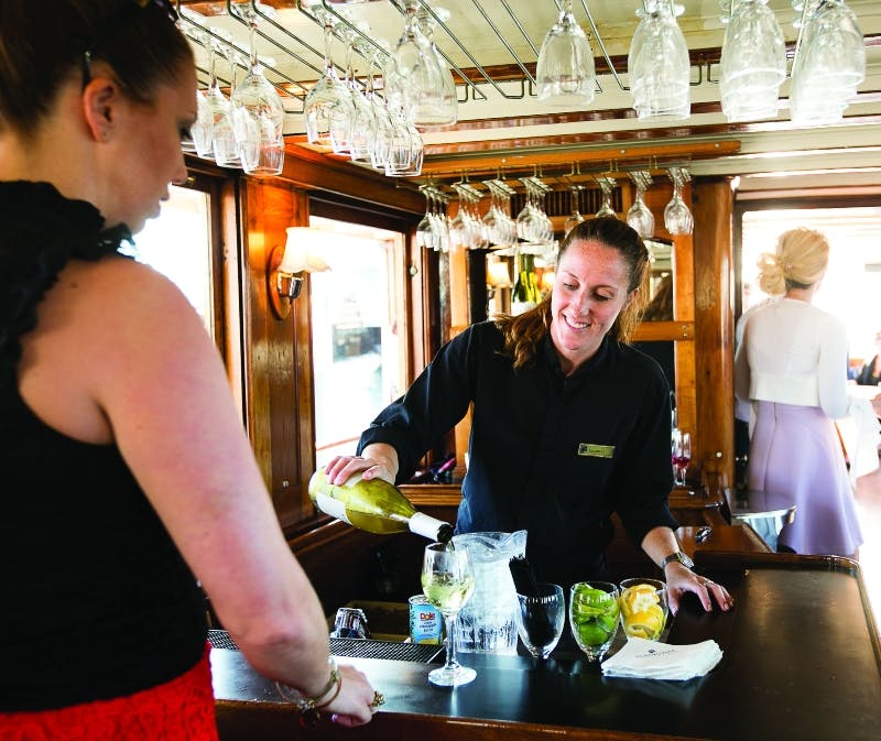 A bartender in a black uniform pours wine for a woman in a black top and red skirt at a bar with hanging glasses.