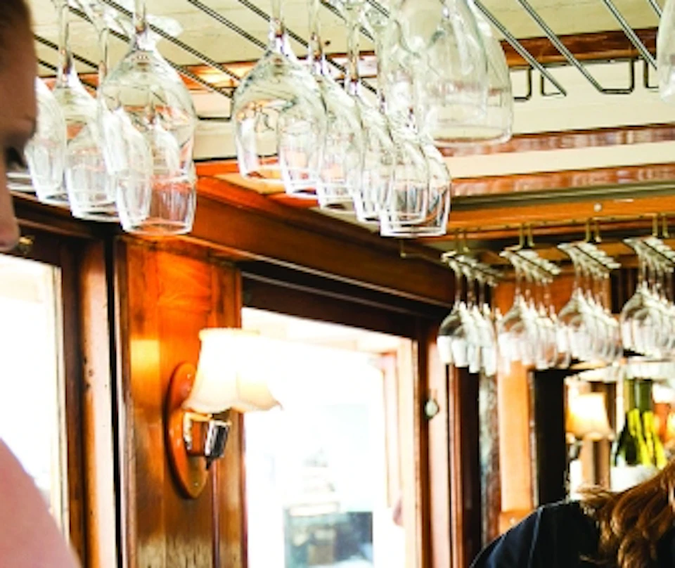A bartender in a black uniform pours wine for a woman in a black top and red skirt at a bar with hanging glasses.