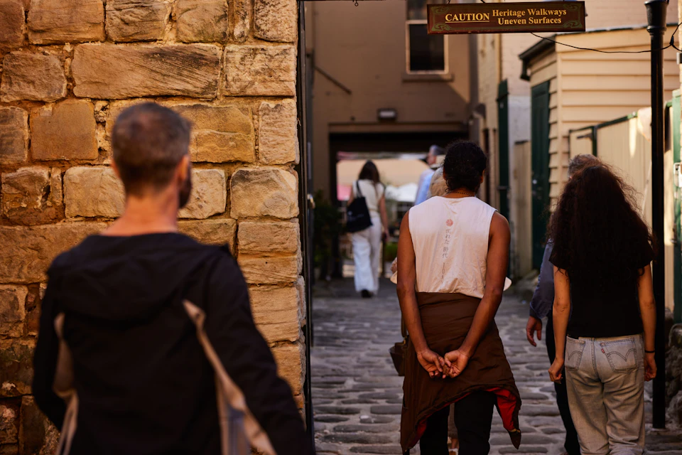 People walking through a narrow stone-paved alley with historic walls, a caution sign above, and buildings in the background.
