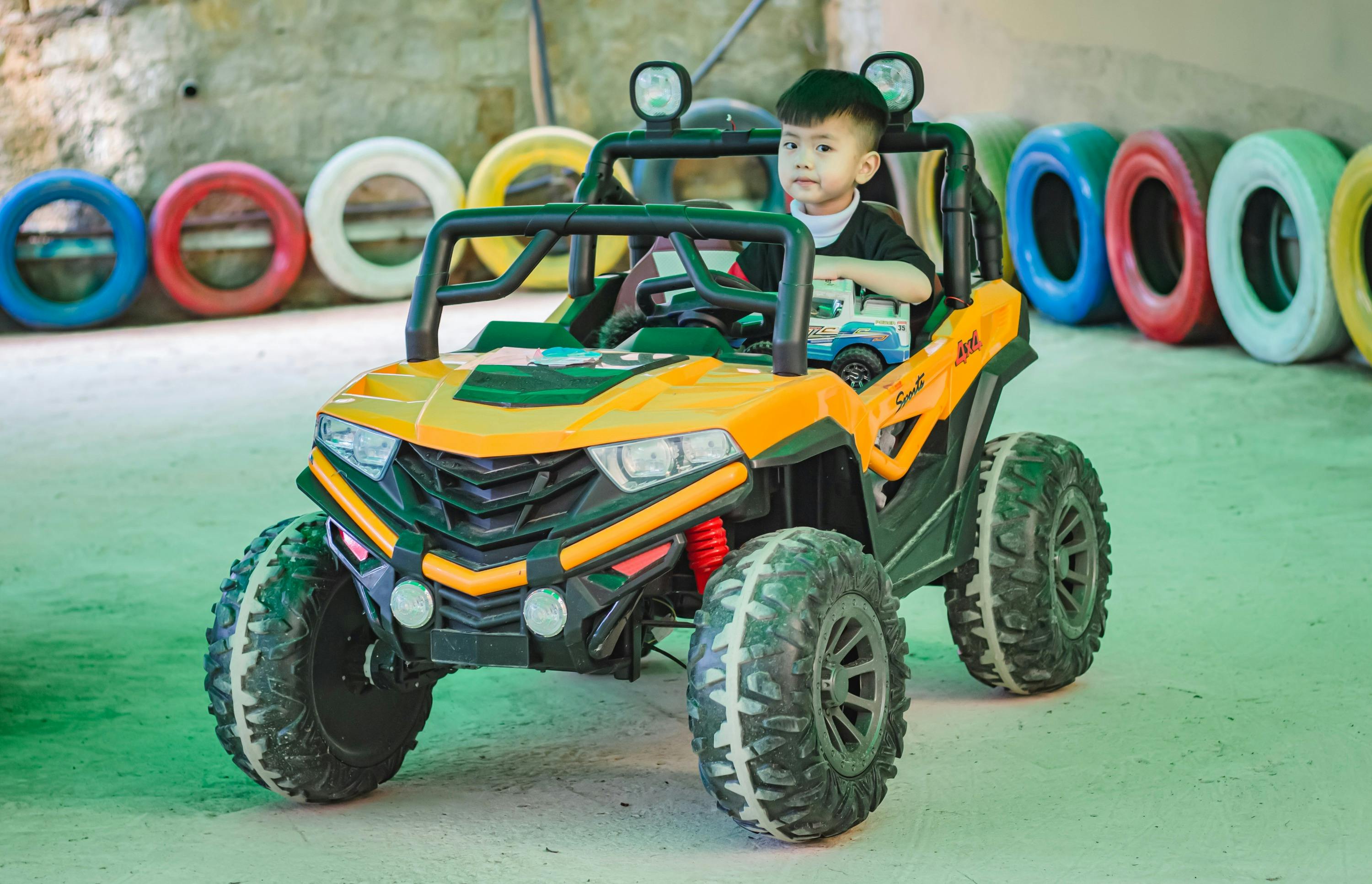 A child sitting in a toy off-road vehicle in an indoor setting with colorful tires in the background.