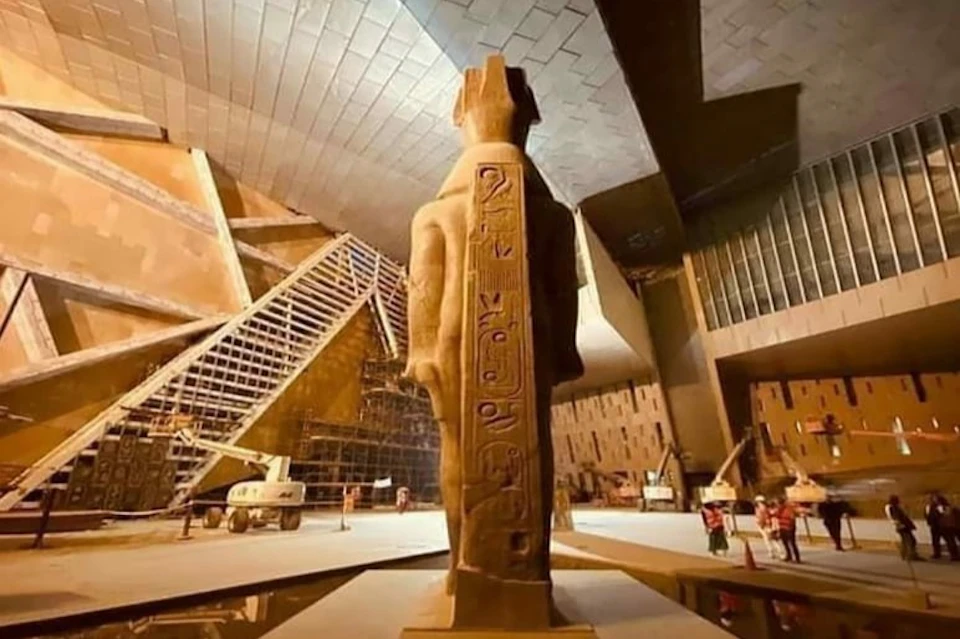 A large statue with hieroglyphs stands inside a modern, angular building under construction, with cranes and scaffolding visible.