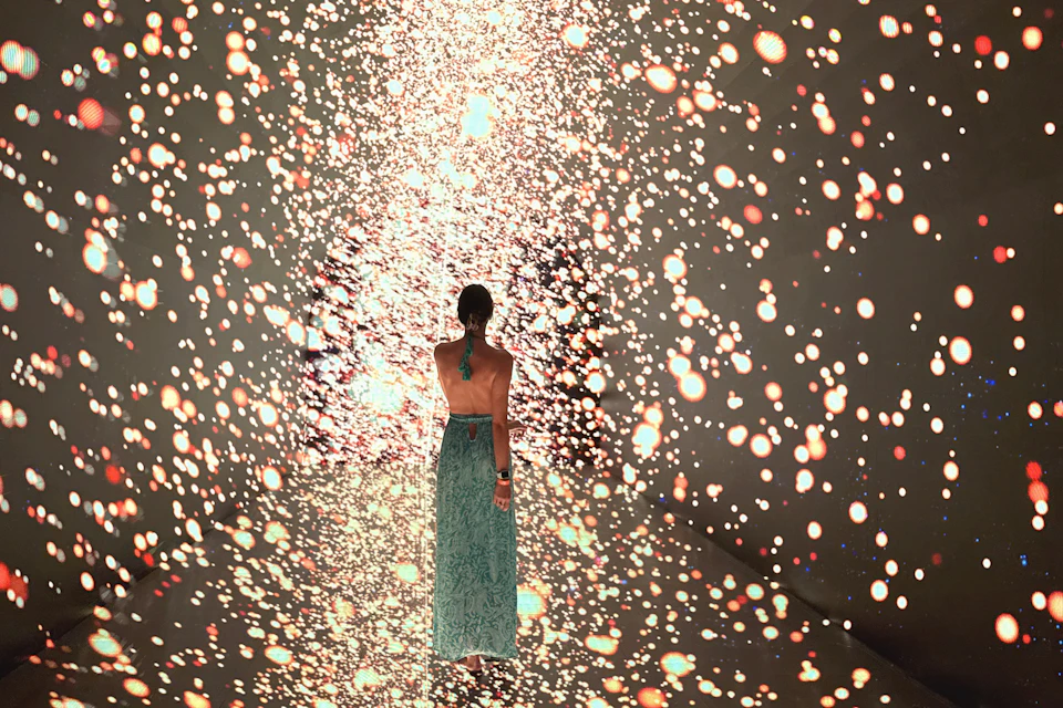 A person in a long, flowing dress stands facing a cascade of glowing lights in an immersive art installation.