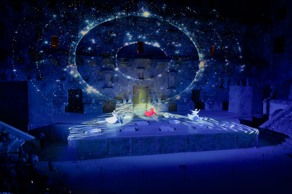 A stage performance with dancers in white and red costumes under a backdrop with a spiral galaxy projection.