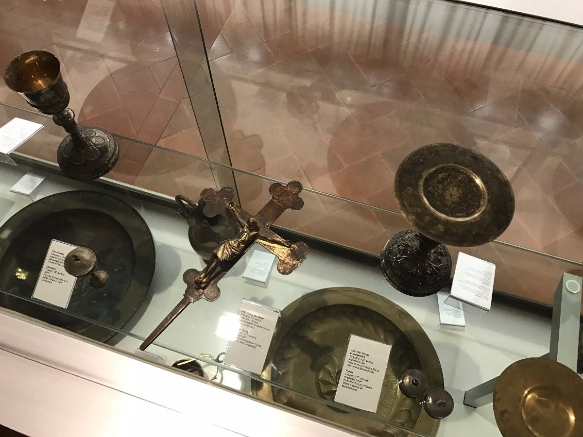 Szklana gablota z metalowymi artefaktami religijnymi, w tym krzyżem, talerzami i świecznikami, z których każdy jest oznaczony kartami opisowymi.