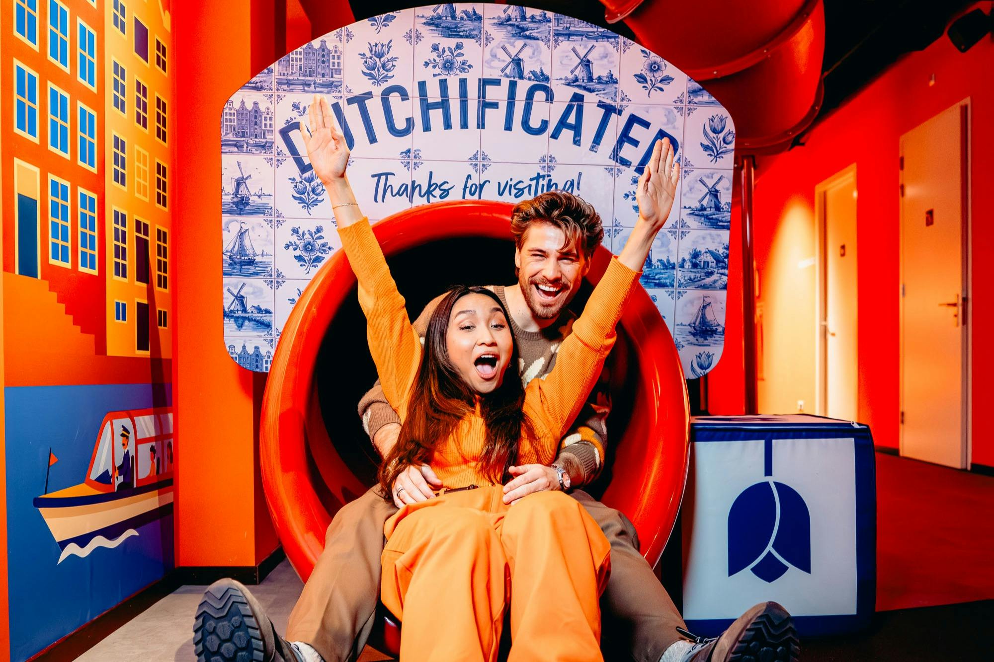 Zwei Personen rutschen eine rote Rutsche hinunter, über der ein Schild mit der Aufschrift "Dutchificated" hängt. Die Szene ist bunt und lebendig.
