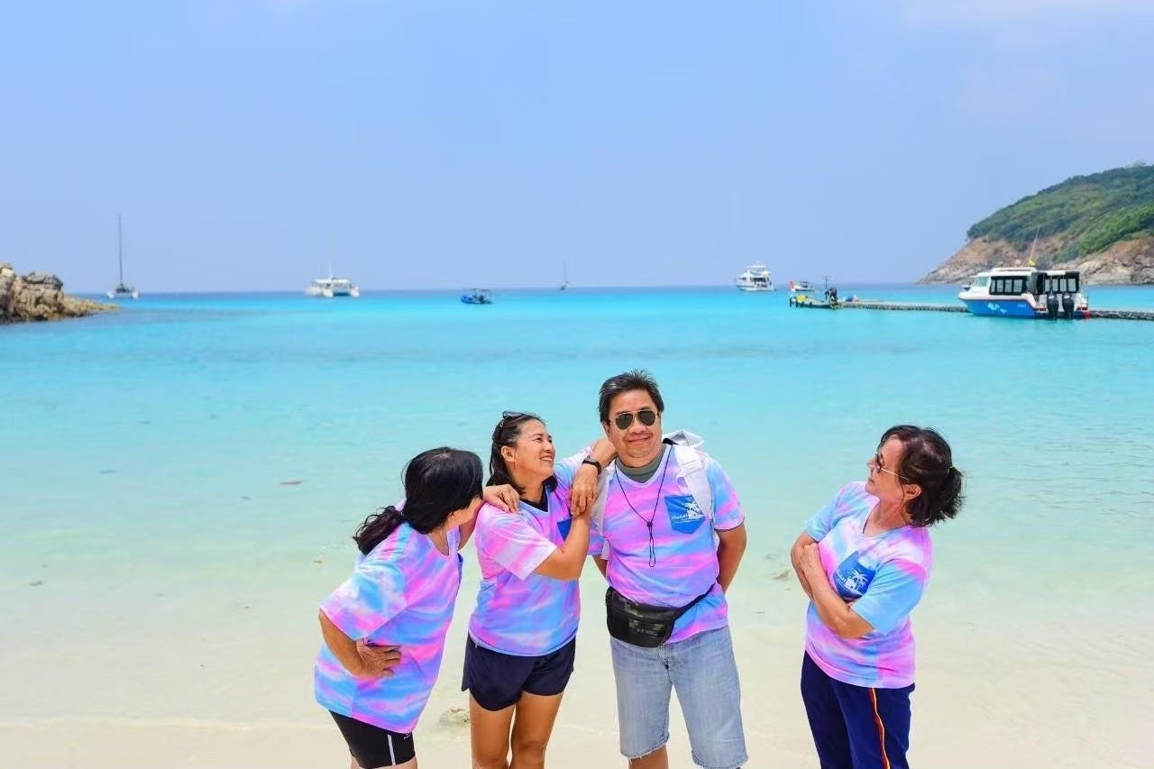 四个穿着匹配扎染衬衫的人在海滩上微笑着摆姿势，背景是清澈的蓝水和船只。