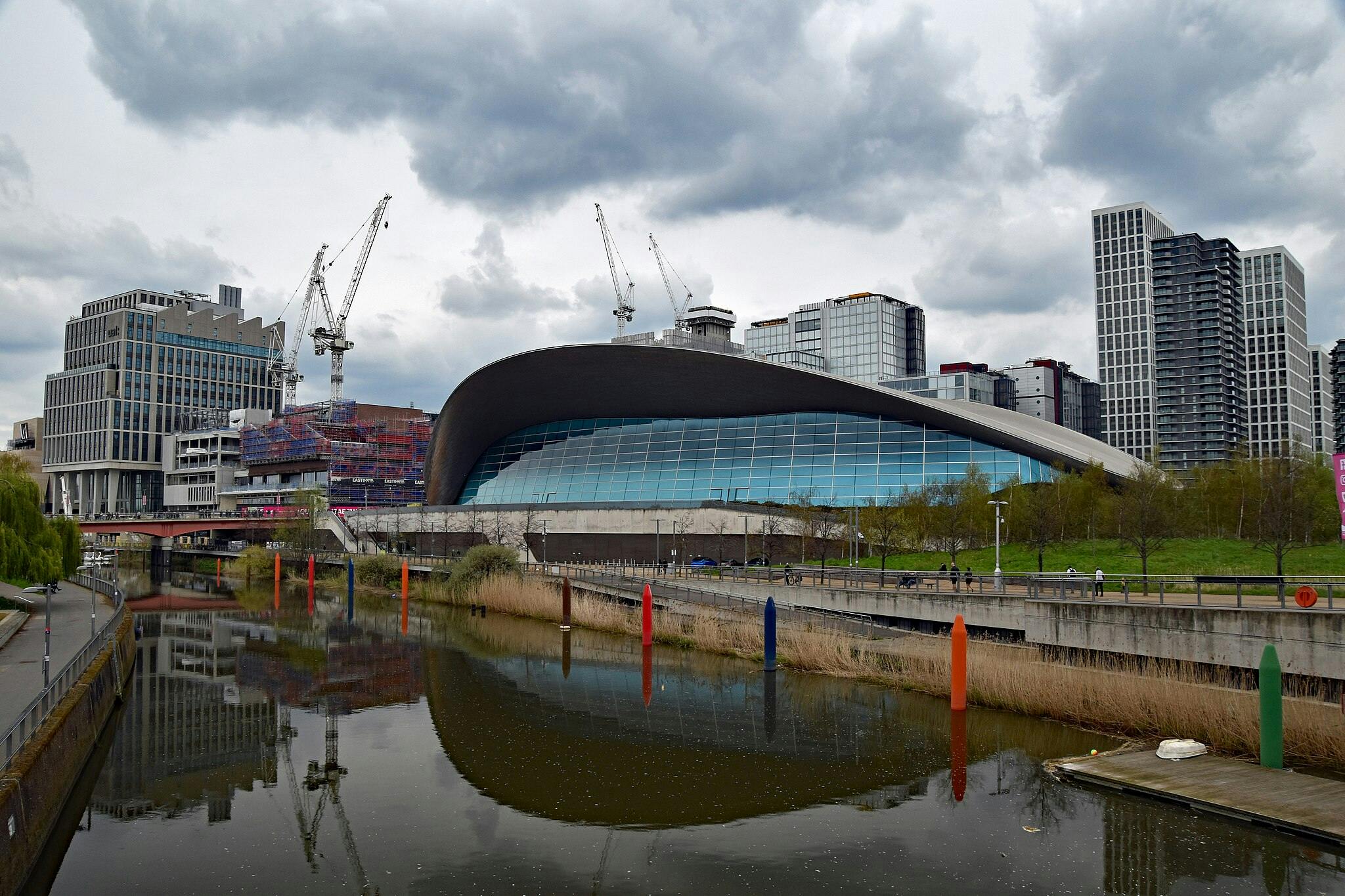 Edifici modern de sostre ondulat amb gran façana de vidre, envoltat d'edificis de gran alçada i grues, reflectit en una via fluvial tranquil·la.