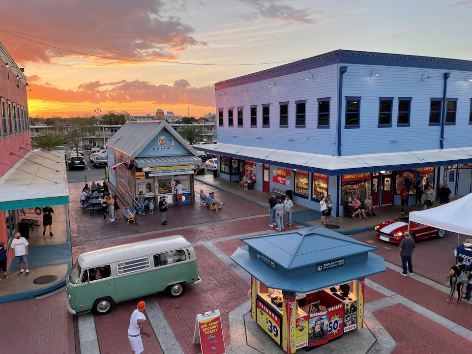 People walking around a retro-themed shopping area at sunset, featuring a food stand, shops, and a green vintage van.