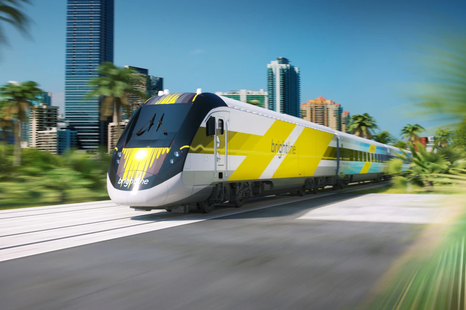A modern yellow and white passenger train traveling through a cityscape with palm trees and tall buildings in the background.