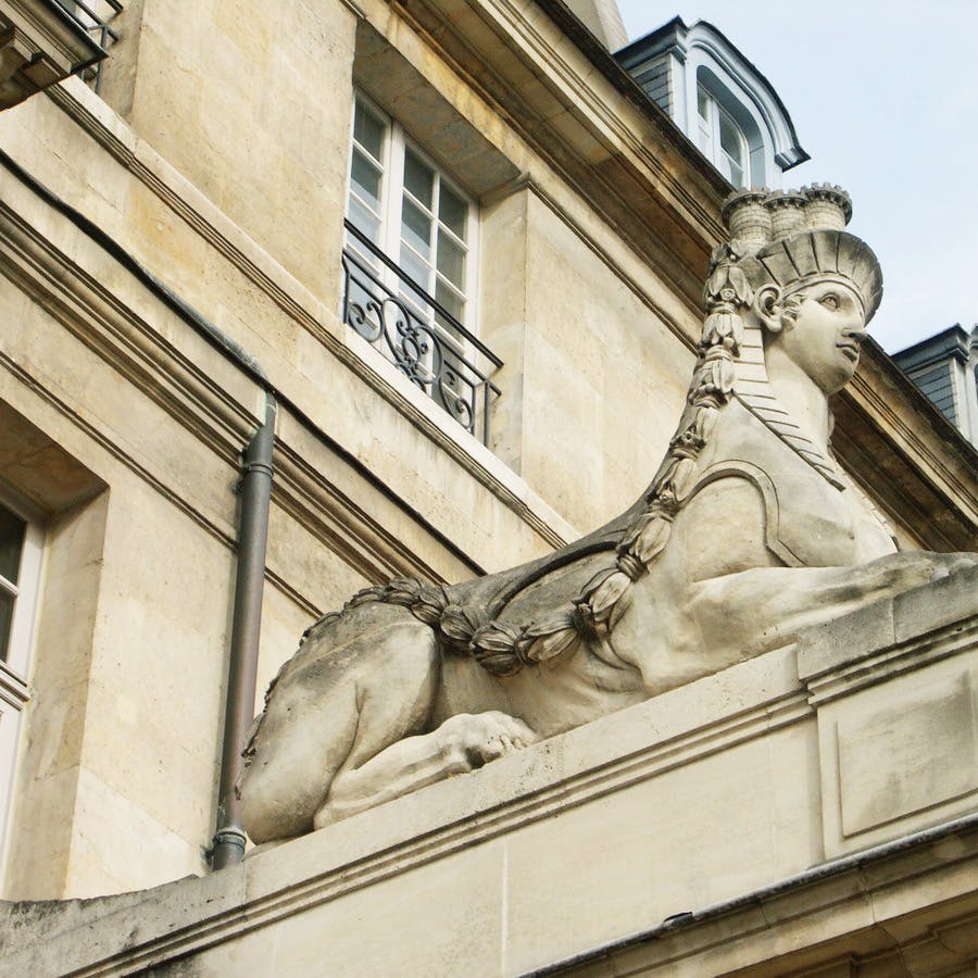 A stone sphinx sculpture is integrated into the façade of a historical building with decorative windows and a steep roof.