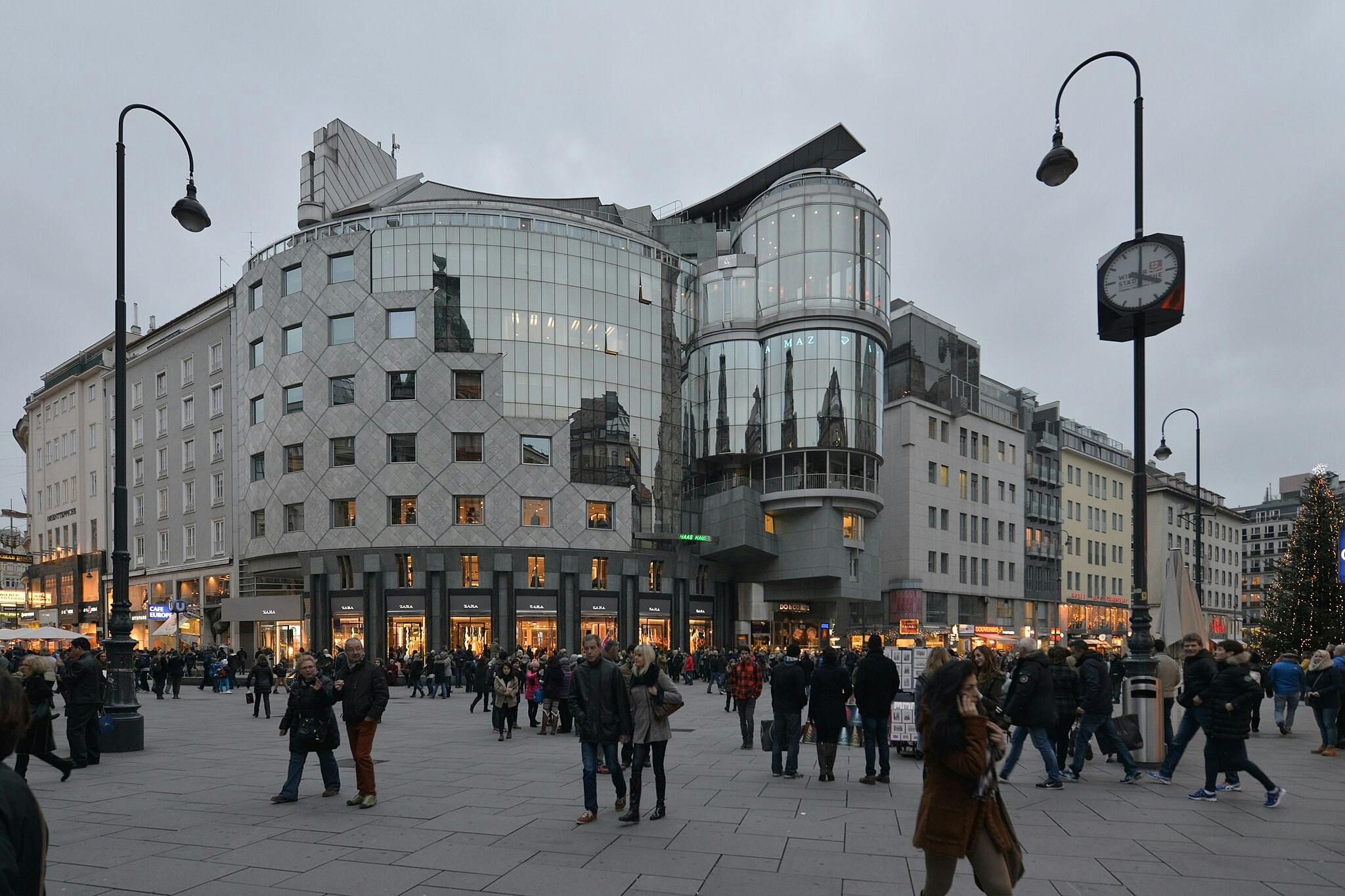Přeplněné náměstí s moderními prosklenými budovami, výkladními skříněmi, pouličními lampami a lidmi, kteří chodí a nakupují.