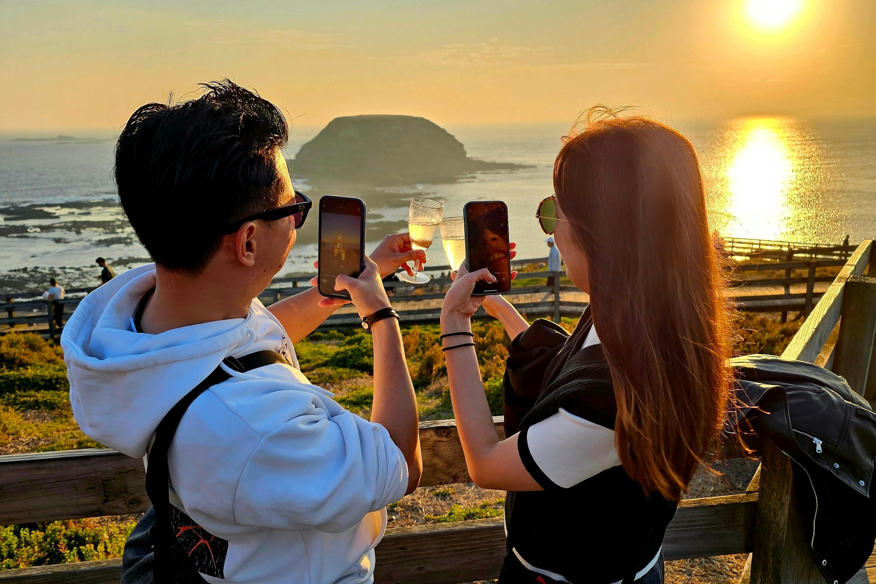 Two people taking photos of a coastal sunset with their phones, one holding a drink, overlooking water and a distant island.