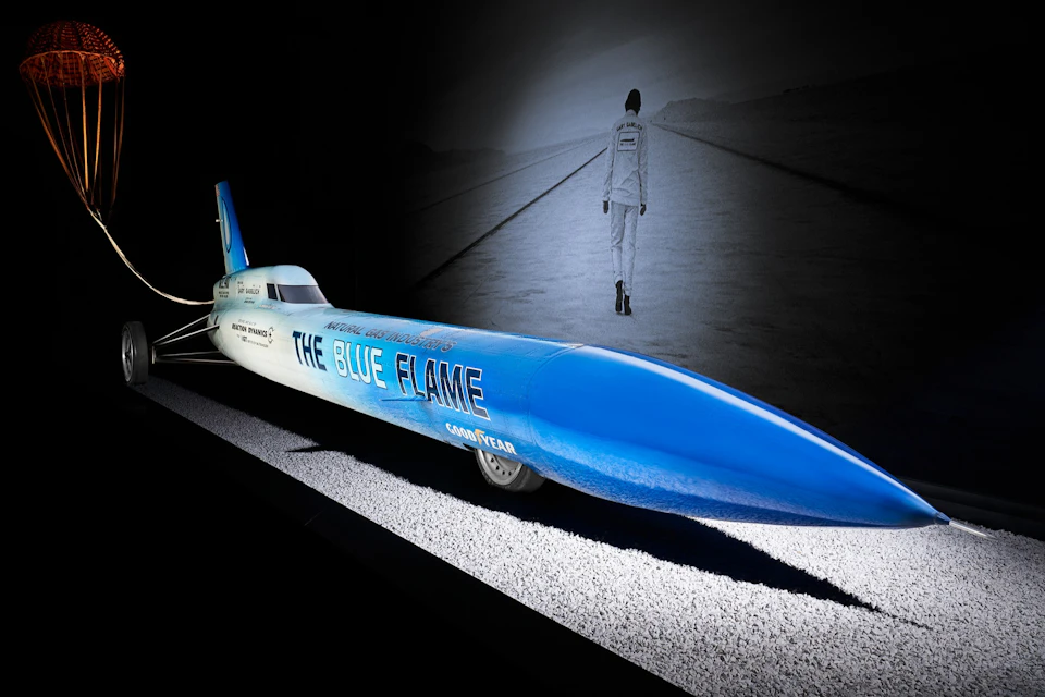 A blue rocket car labeled "The Blue Flame" on display, with a black-and-white mural of a person on a road in the background.