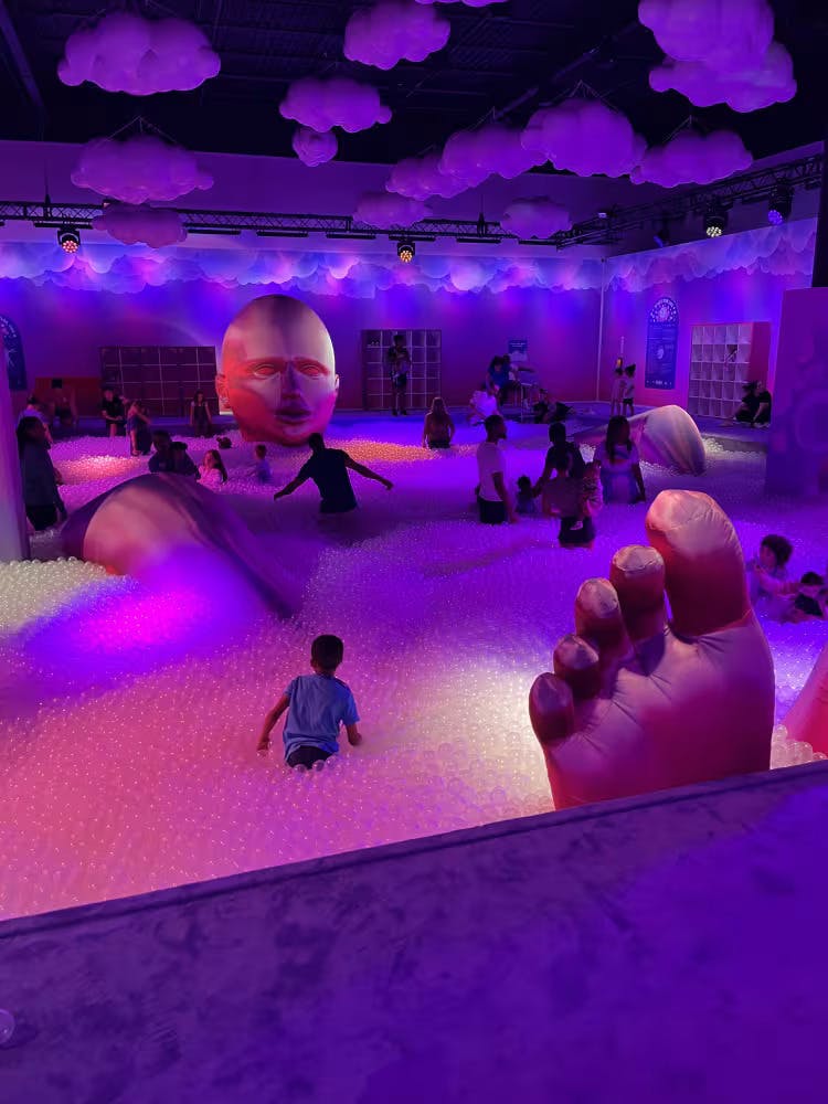 孩子们在灯火通明的球坑里玩耍，头部、手部和脚部的大型雕塑被漂浮的云彩装饰所包围。