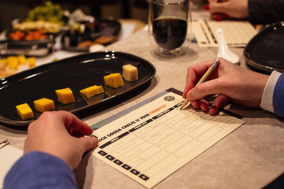 Person writing on a tasting sheet with a pencil, next to a plate of cheese samples and a glass of dark beverage.