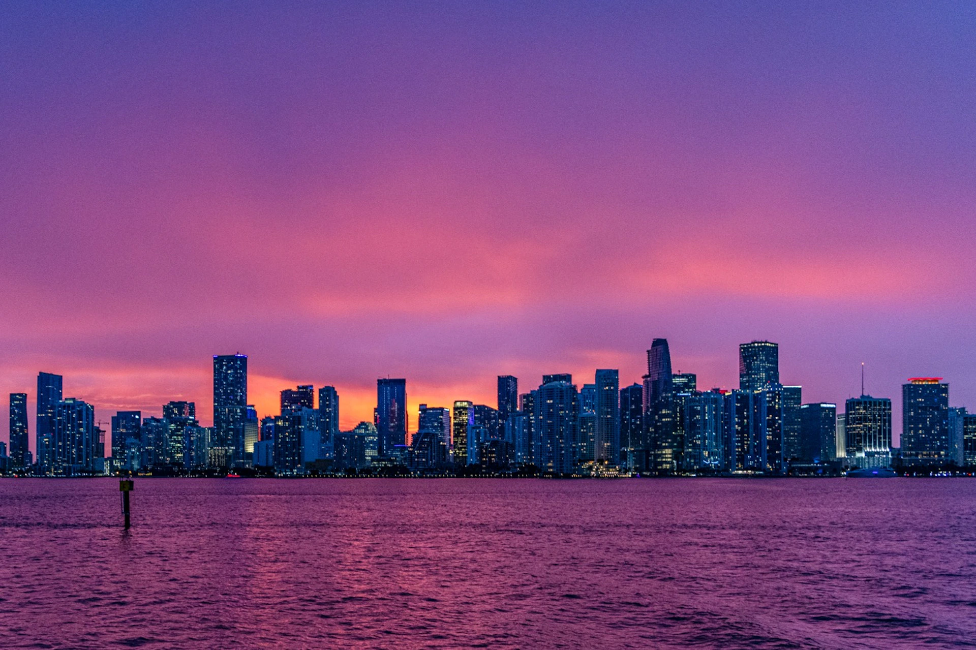 City skyline at dusk with illuminated buildings under a vibrant sunset sky, viewed across a calm body of water.
