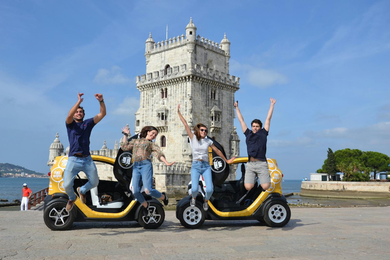 Four people jumping with joy beside two small yellow vehicles in front of a historic tower on a clear day.