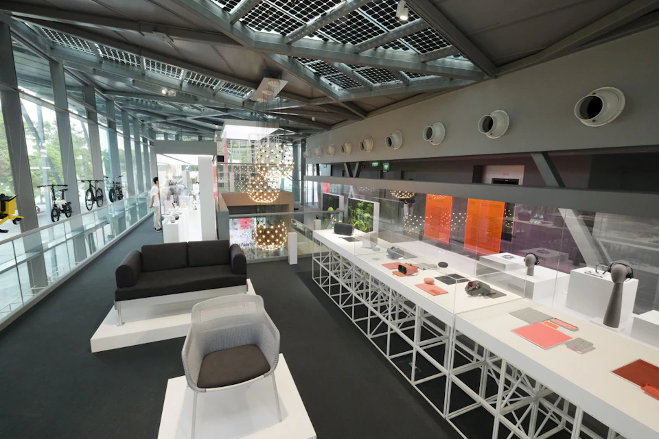 A modern showroom with various design items, including a couch, chair, and gadgets displayed on white tables and shelves.