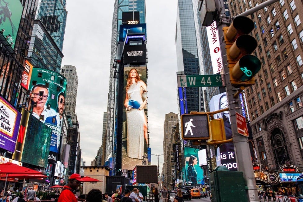 Rušná městská scéna s vysokými budovami a barevnými digitálními billboardy, včetně velké reklamy na ženu v bílých šatech.