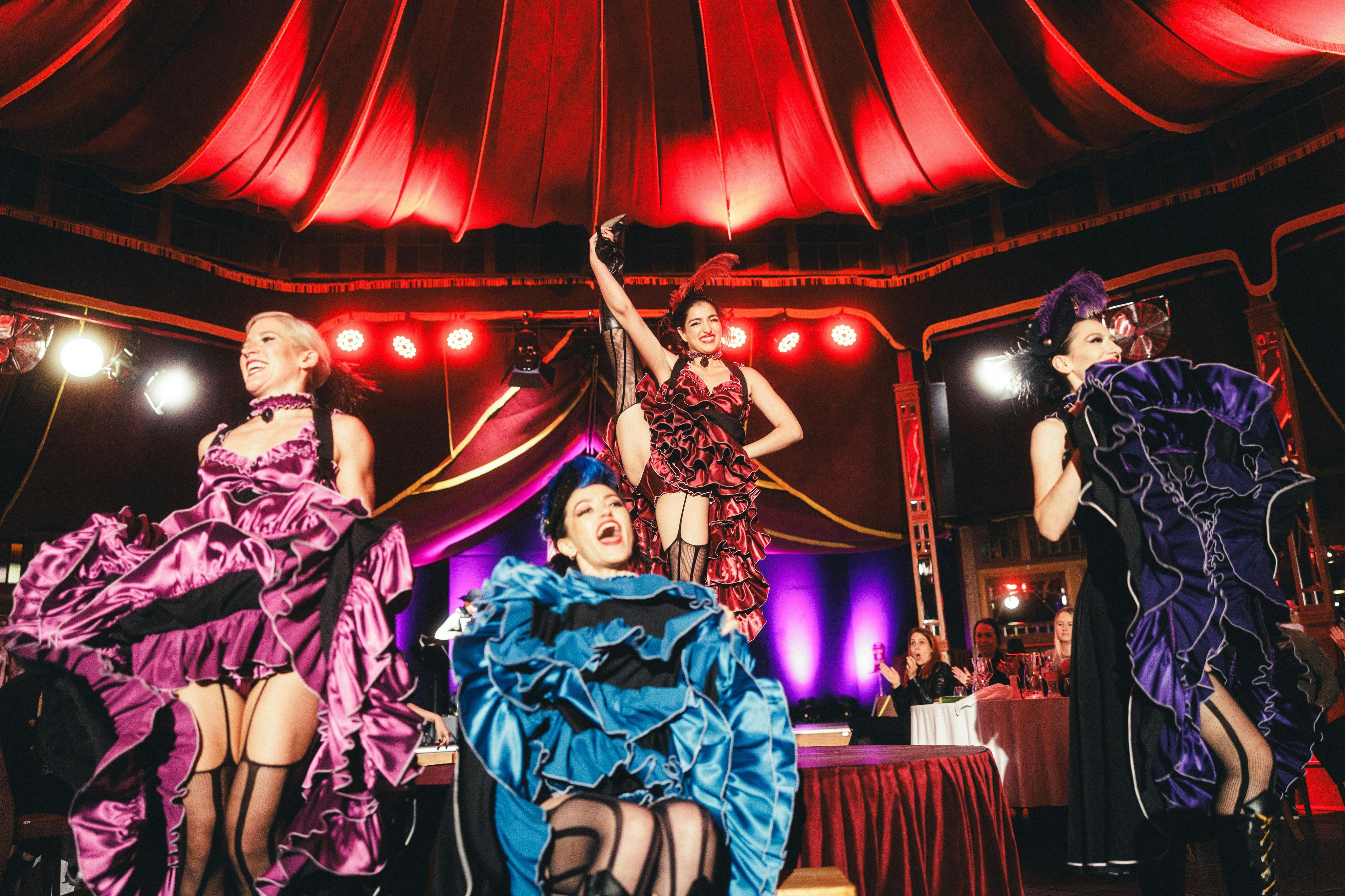 Four women in colorful dresses and stockings perform a lively can-can dance on a lit stage under a red canopy.
