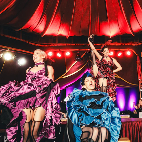 Fyra kvinnor i färgglada klänningar och strumpor utför en livlig can-can-dans på en upplyst scen under ett rött tak.
