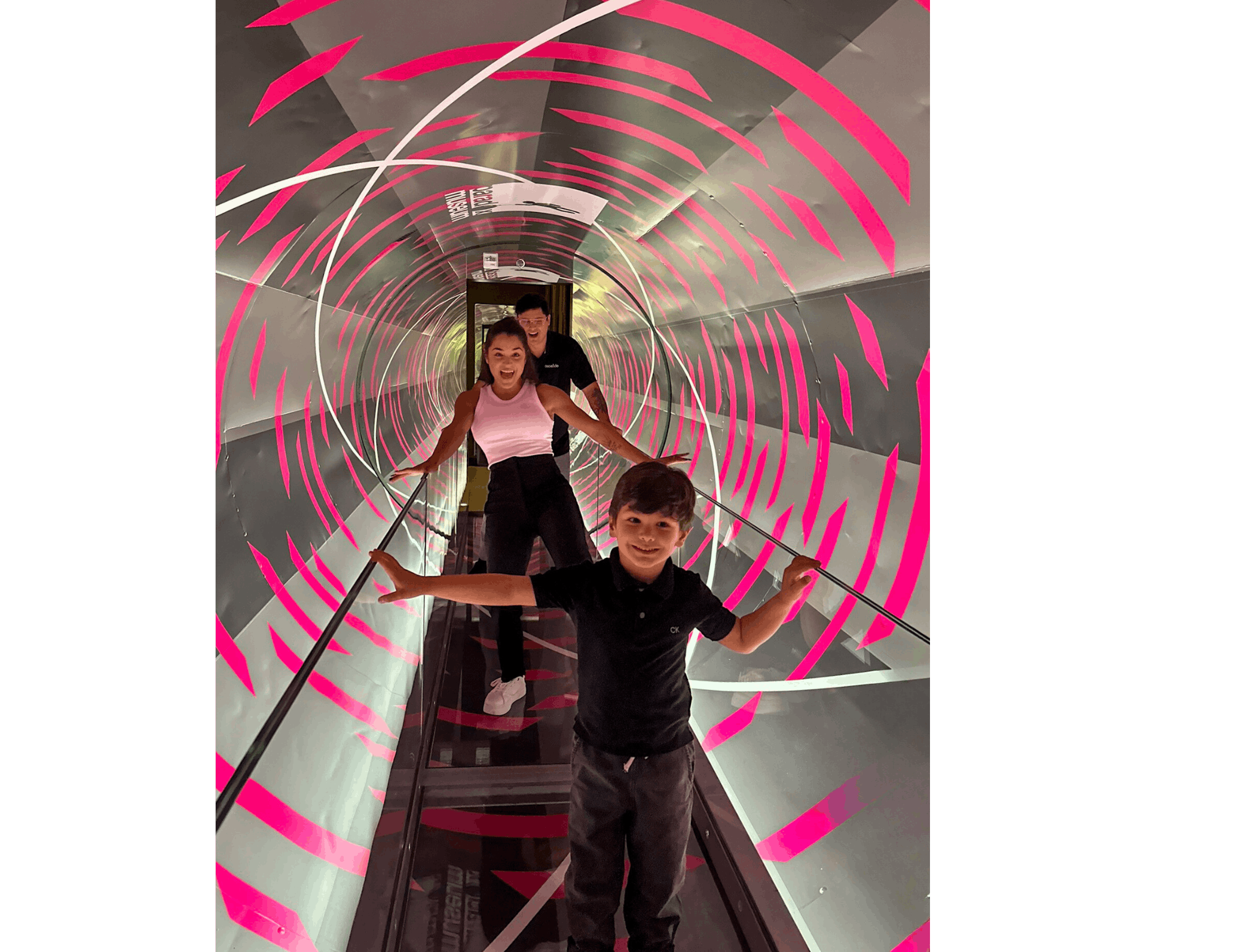 Trois personnes marchent dans un tunnel tournant avec des rayures roses et blanches, en se tenant aux rampes pour garder l'équilibre.