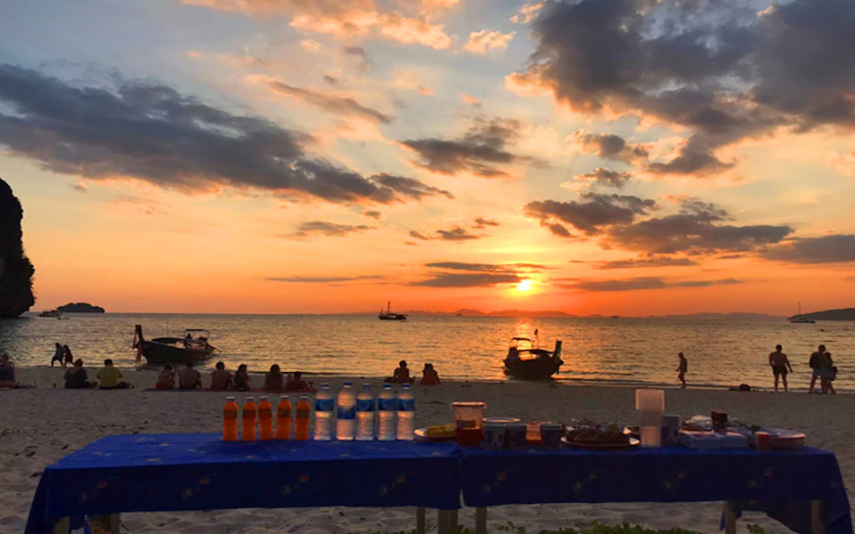 Sunset at the beach with boats on water, people sitting on the sand, and a table set with drinks and food in the foreground.