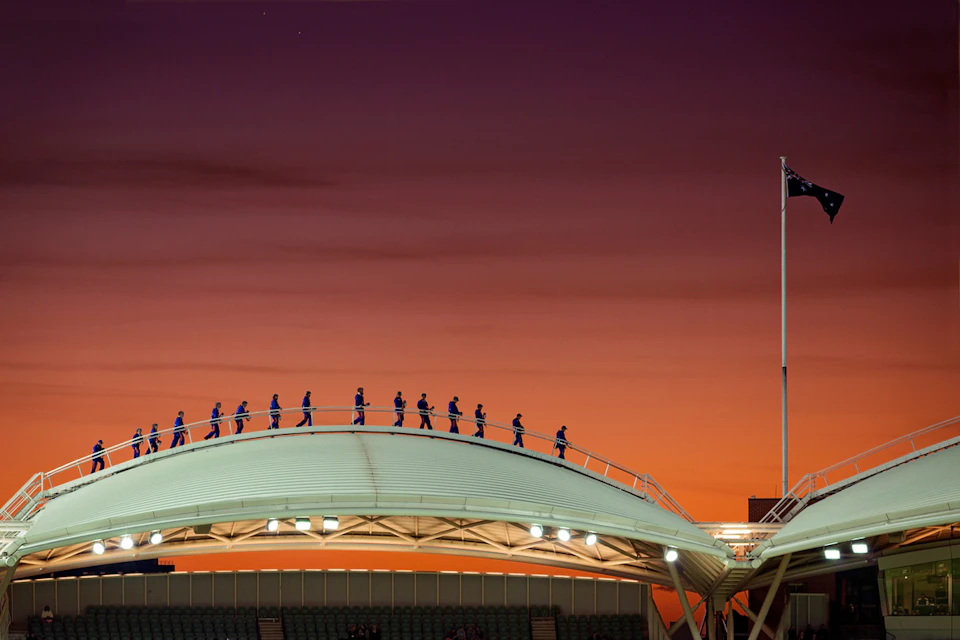 People walking on a curved roof structure at sunset with a flagpole on the right side.