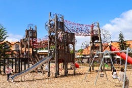 A colorful outdoor playground with climbing structures, slides, rope bridges, and swings under a clear blue sky.