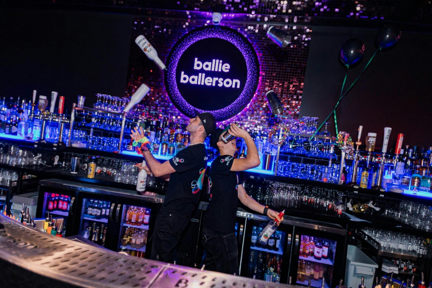 Two bartenders perform flair techniques with bottles and shakers in a bar with a neon 'Ballie Ballerson' sign.