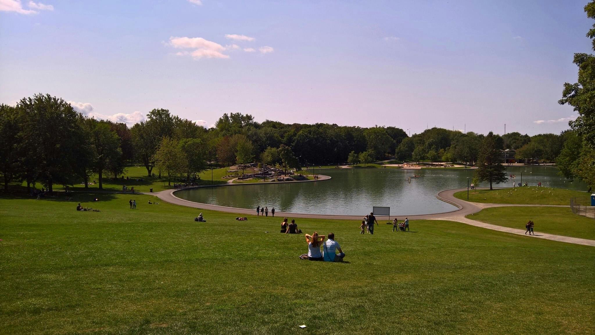 People scattered on a grassy hill overlooking a lake, with trees in the background and a blue sky above. Paths wind around the water.