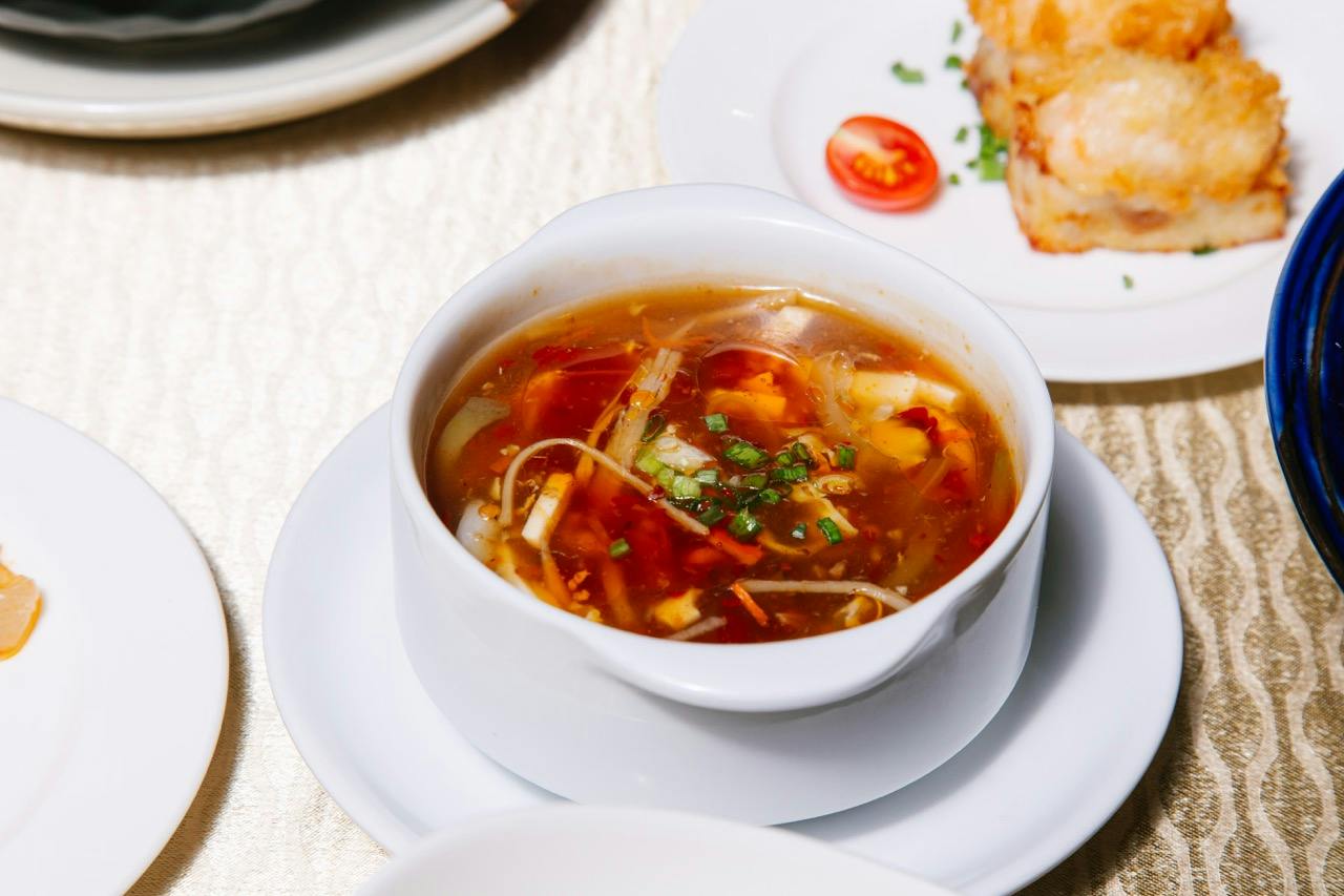 A bowl of soup with vegetables and herbs, served on a white plate. A tomato garnish and bread are visible in the background.