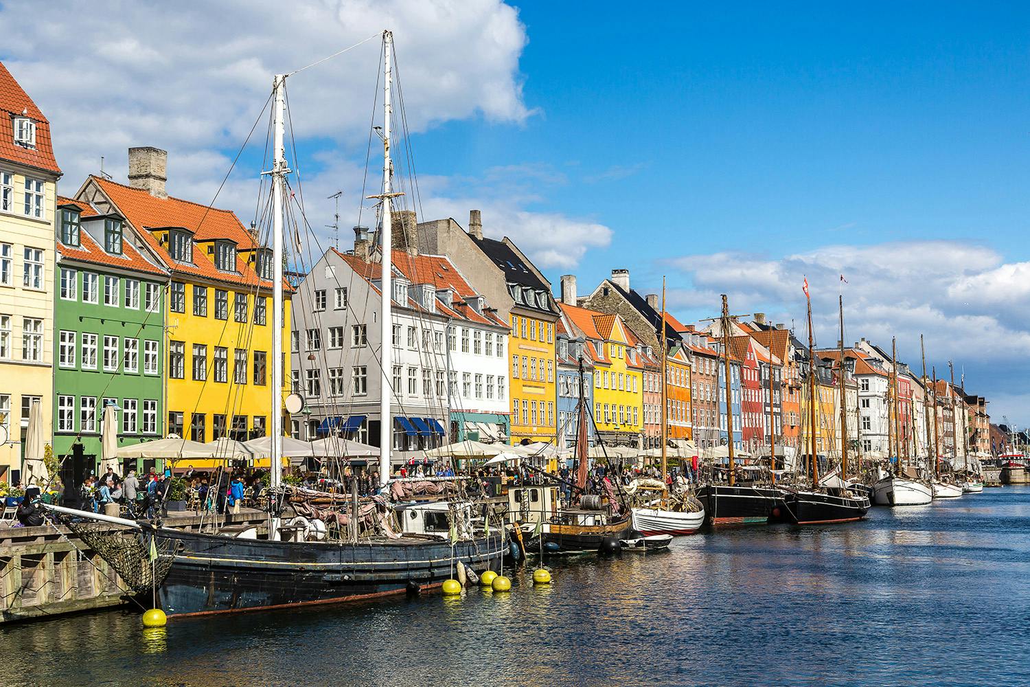 Colorful buildings, boats docked along a canal, and outdoor cafes under a sunny sky with scattered clouds.