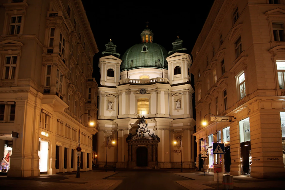 Illuminated baroque church with a green dome, flanked by buildings on a narrow street at night.