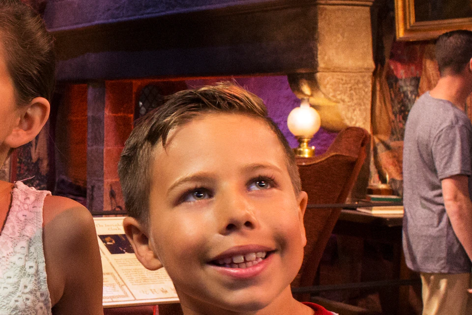 Two children smile inside a room with a medieval theme, featuring paintings, a chandelier, and people in the background.