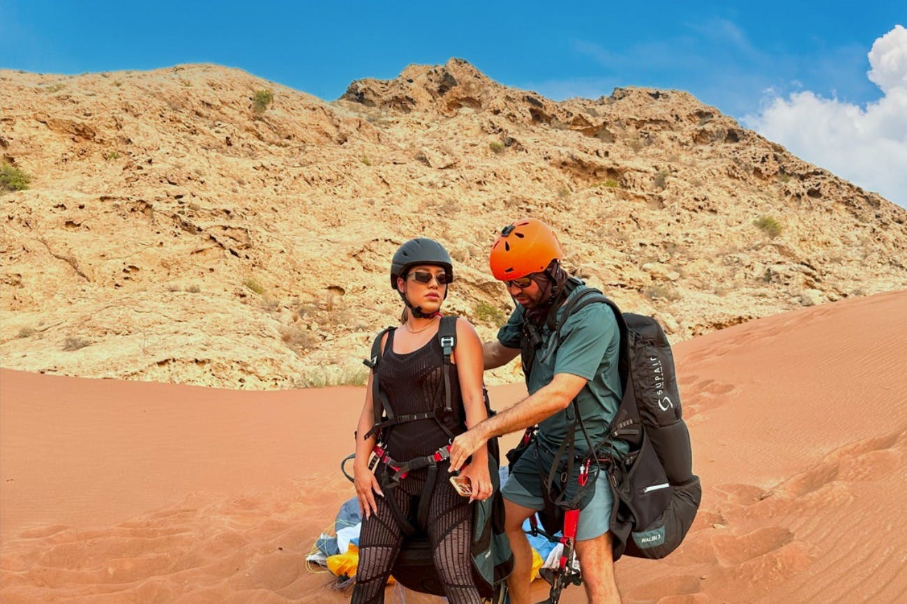 Ένας άνδρας με πράσινη στολή βοηθά μια γυναίκα που φοράει αθλητικό εξοπλισμό και κράνος να προσαρμόσει την εξάρτυσή της σε ένα σκηνικό στην έρημο.