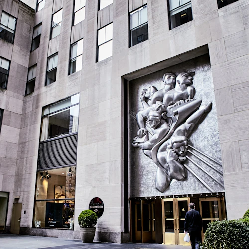 Großes Steingebäude mit hohen Fenstern, einer Reliefskulptur aus Metall, Eingangstüren mit der Aufschrift "Rockefeller Plaza" und einer Beschilderung der Bank of America.