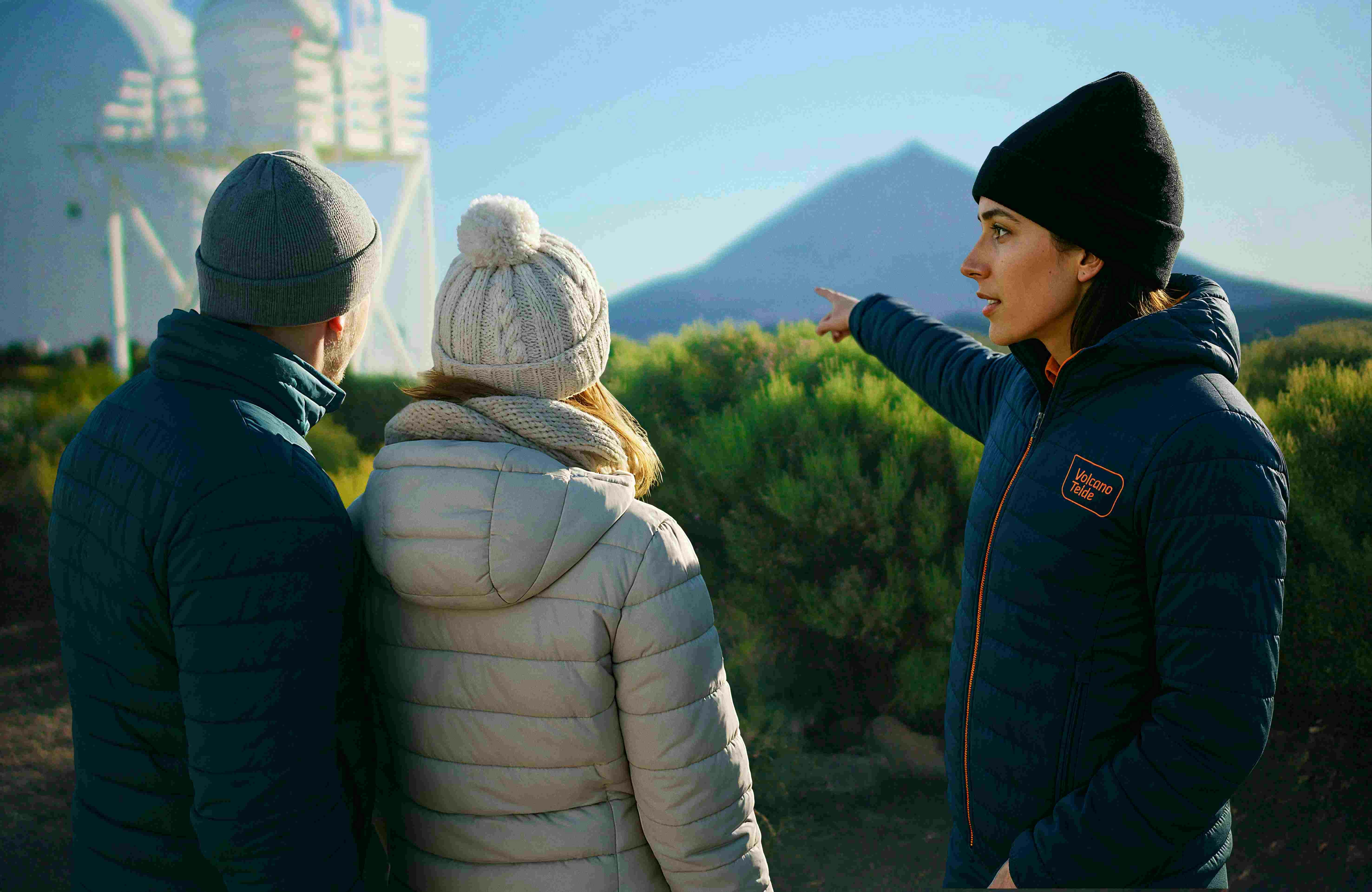 Two people in winter jackets and hats stand outdoors. One points towards a distant mountain.