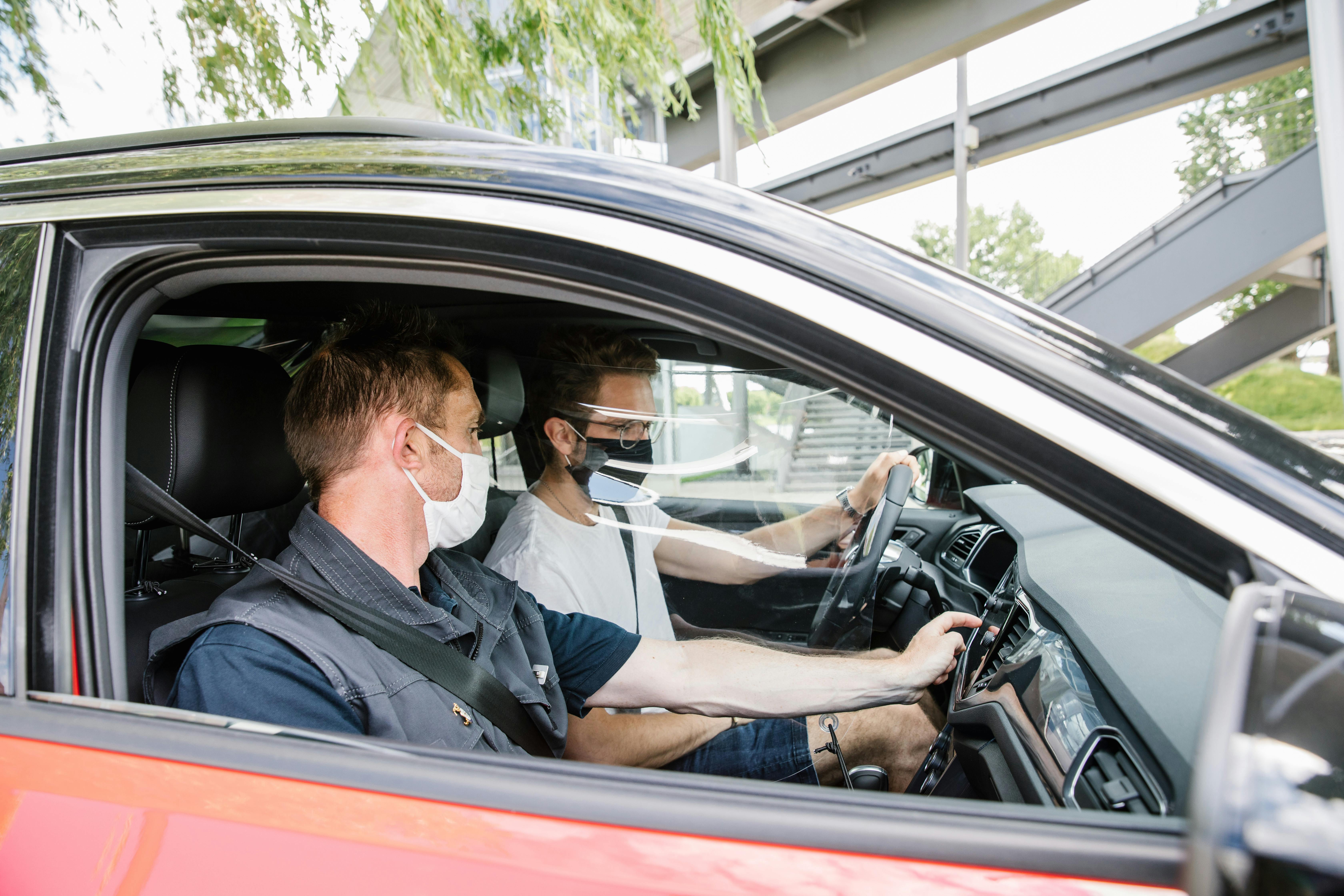 Two men wearing face masks in a car. The driver is adjusting the controls while the passenger looks on attentively.