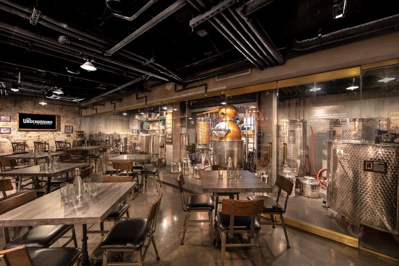 Industrial-style distillery seen through glass from a seating area with wooden tables and chairs. Copper stills and steel tanks are visible.