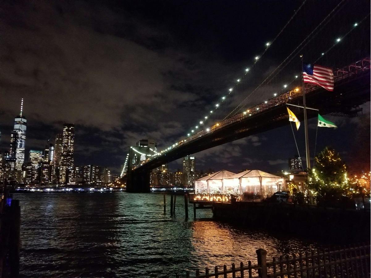 Nighttime view of a lit-up bridge over water with city lights, a waterfront venue, and flags in the foreground.