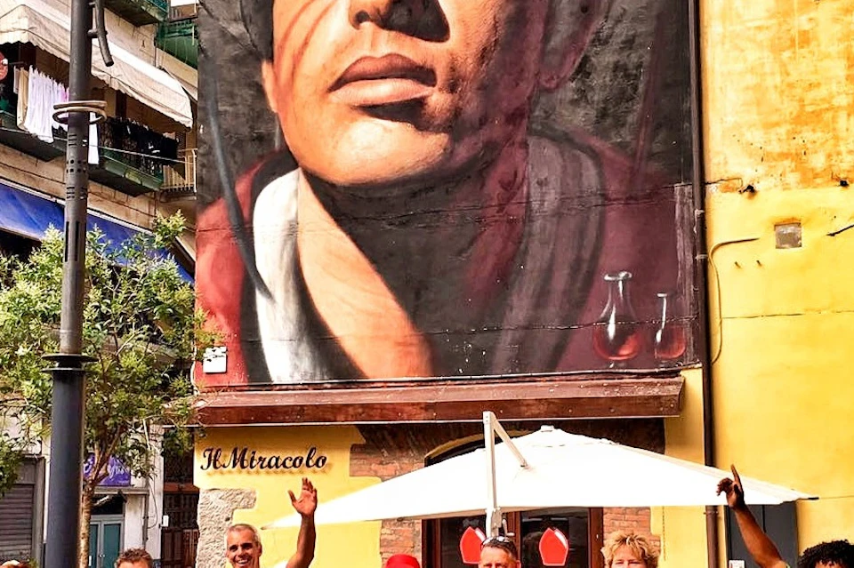 A diverse group of people pose with bicycles under a large mural of a crowned man on a city street.