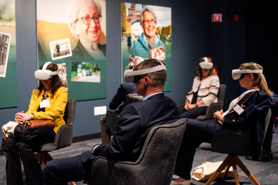 People seated in a room wearing virtual reality headsets, with large portraits displayed on the wall behind them.