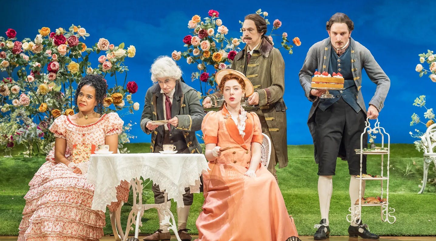 Ομάδα ανθρώπων με κοστούμια εποχής που κάνουν ένα πάρτι τσαγιού σε εξωτερικούς χώρους, με φλοράλ φόντο και ένα άτομο που σερβίρει τούρτα.