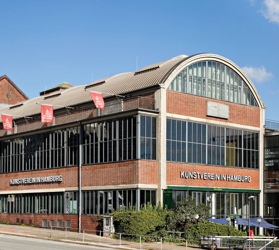 A large brick and glass building with "Kunstverein in Hamburg" written on it, featuring red banners and a curved roof.