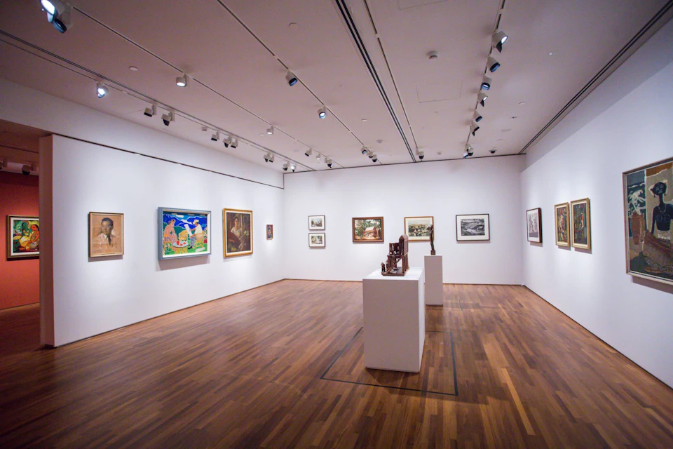 A modern art gallery with wooden flooring, white walls, various paintings, and sculptures on white pedestals.