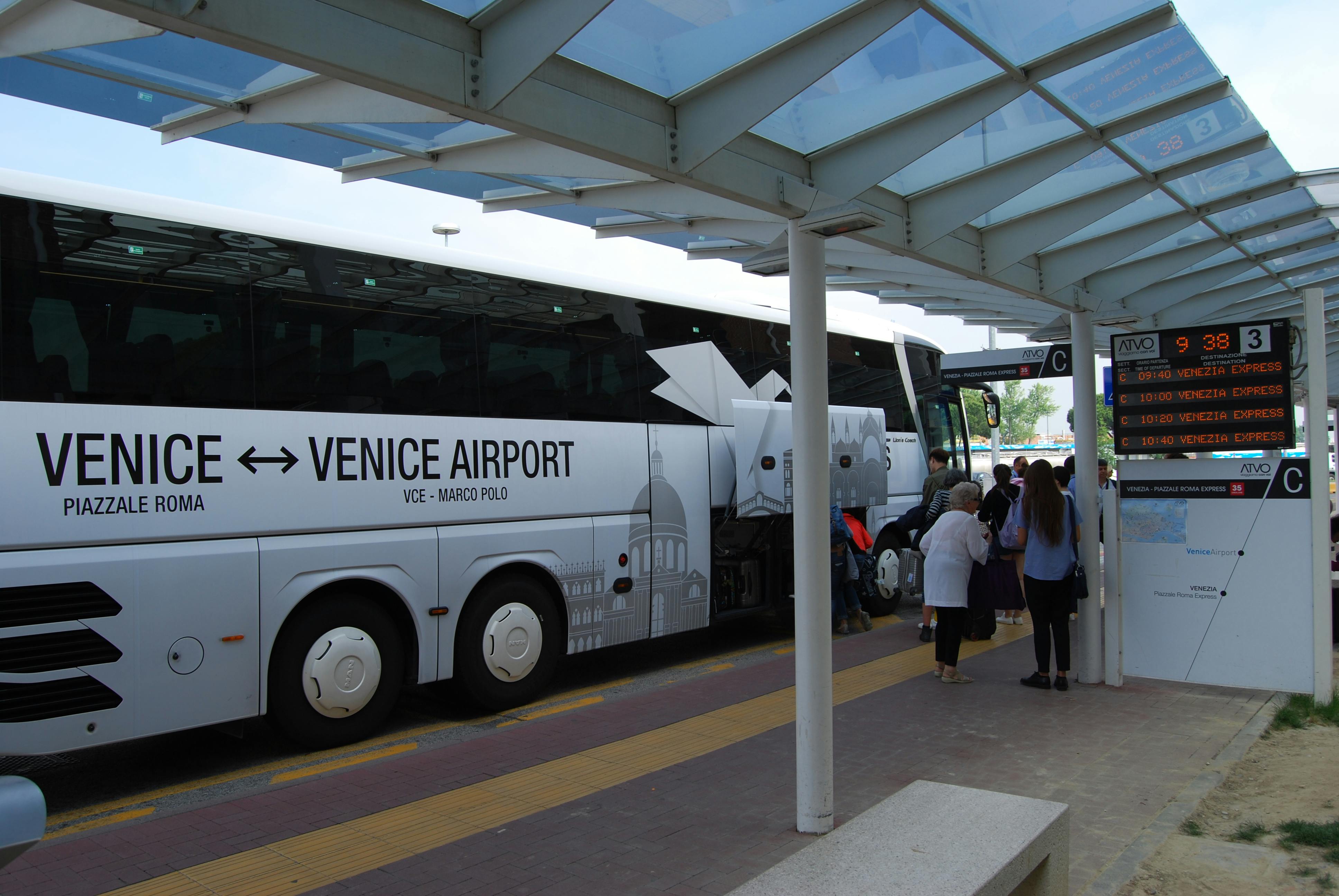 People boarding a bus labeled "Venice Airport" under a glass canopy at a bus terminal.