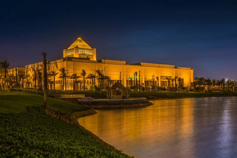 Illuminated building with a glass pyramid roof, surrounded by palm trees and water, under a deep blue sky at night.