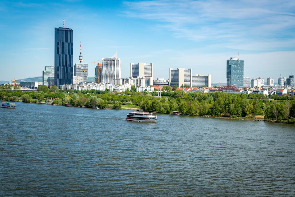 City skyline with modern high-rise buildings, a boat on a river, and trees lining the riverbank under a clear blue sky.